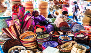 Colorful handcrafted baskets and mats displayed at a Kenyan market, with vendors and shoppers in the background.