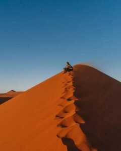 A person sitting on top of a tall sand dune with a trail of footprints leading up, under a clear blue sky.